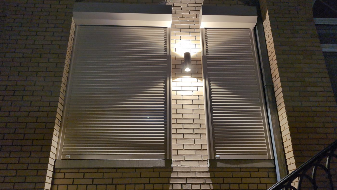 Two closed aluminum security shutters on a brick home facade at night